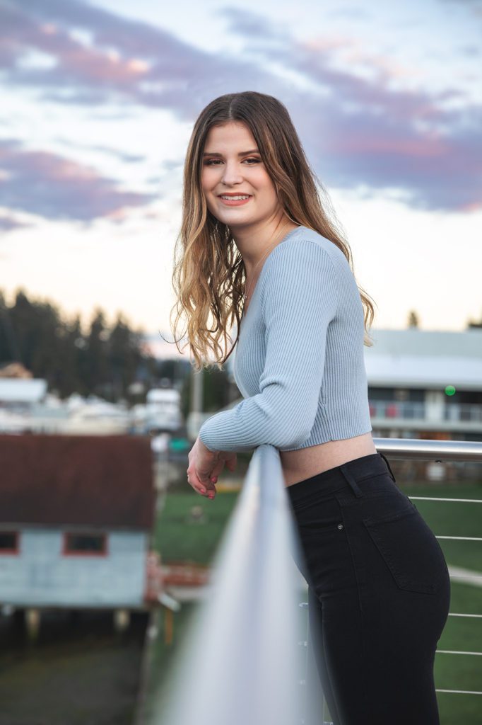 high school girl leaning over railing in grey top and black jeans Skansie brother's Park and netshed downtown gig harbor washington. 