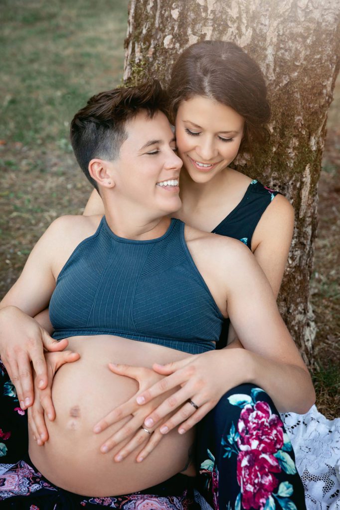 Romantic lesbian couple posed by tree for maternity session at Scenic Beach Sate Park Seabeck Washington. 